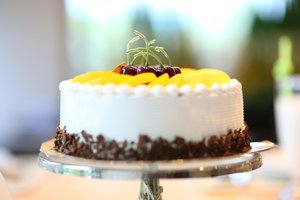 A round white frosted cake topped with yellow peach slices and cherries, garnished with chocolate shavings around the base, placed on a glass cake stand.
