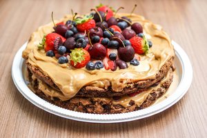 A layered chocolate cake with creamy caramel frosting, topped with fresh strawberries, cherries, and blueberries, displayed on a white plate against a wooden background.