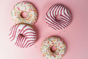 Four donuts on a pink background: two with white icing and colorful sprinkles, and two with white icing and pink stripes.