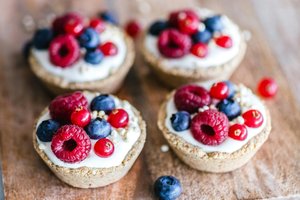 Four mini tartlets on a wooden surface, topped with white cream, fresh raspberries, blueberries, and red currants, garnished with seeds.
