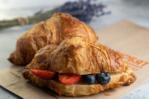Two flaky croissants are placed on a brown paper bag. One is filled with slices of fresh strawberries and blueberries. In the background, a bunch of lavender lies slightly out of focus.